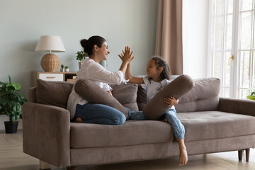 Happy mom and cheerful little kid giving high five after pillow fighting battle, sitting on couch, holding cushions, clapping hands, laughing, talking, playing active games. Family leisure concept