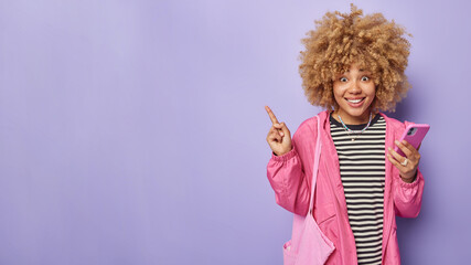 Horizontal shot of cheerful young woman with curly blonde hair wears striped jumper and pink jacket carries fabric bag uses smartphone points away on blank space isolated over purple background