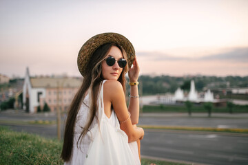 Portrait of a relaxed happy woman in white dress with hat looking at the camera. Cityscape in the background - 520354484