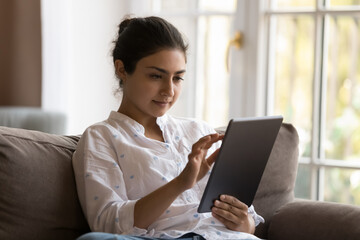 Focused young Indian tablet user woman relaxing on sofa at home, reading book online, shopping on Internet, using app for distant work, touching touchscreen, enjoying leisure, comfort
