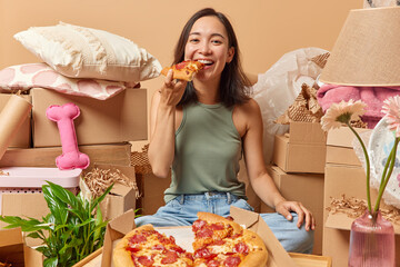 Positive young woman with eastern appearance eats tasty pizza relocates to new apartment enjoys...