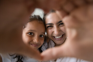 Happy pretty Indian kid girl and excited cheerful mom looking through hand heart frame at camera, smiling, laughing, joining fingers, showing symbol of love. Family, motherhood concept. Close up