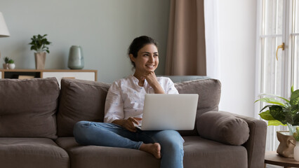 Happy pretty Indian laptop user girl working at laptop from home, thinking over online project, success, achieve, touching chin, looking at window away, smiling, resting on couch, enjoying leisure