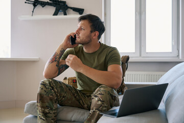 Soldier sitting near laptop on couch and talking on phone