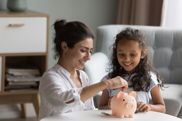 Happy excited Indian mother and kid dropping cash into piggybank. Caring mom teaching kid to save, invest money, collecting coins in piggy bank. Family savings, financial education concept