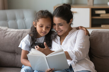 Happy Indian mom hugging little kid on couch, showing smartphone screen to girl on couch, using...