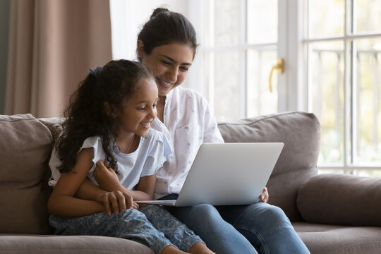 Happy young Indian mother and sweet curious kid watching cartoon movie, series, reading book online, talking on video call, using online learning app on laptop, enjoying leisure