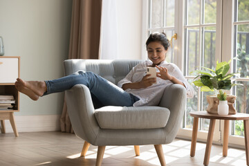 Joyful pretty millennial Indian cellphone user girl resting in armchair at cozy home, touching...
