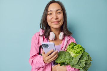 Pleased Asian woman leads healthy lifestyle bought fresh green vegetables keeps to diet holds smartphone downloads app to control eaten calories dressed in pink windbreaker isolated on blue background