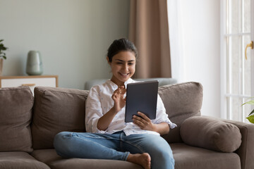 Happy millennial Indian girl using online app on digital device at home, reading book, ebook on...