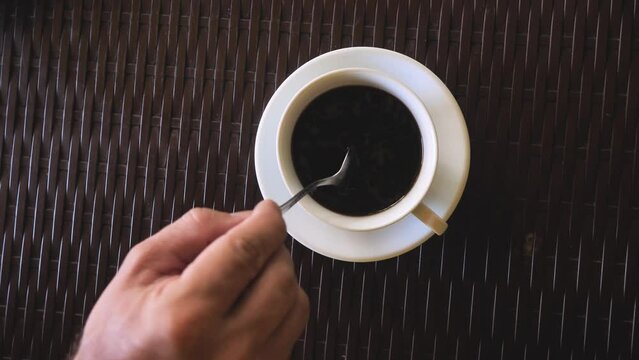 On a dark coffee-colored countertop there is a saucer with a bowl filled with a coffee drink. The drink is dissolved and stirred with a voluntary movement with a spoon. Morning espresso coffee.