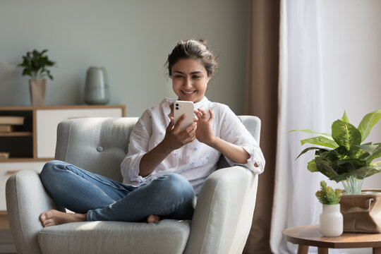 Happy pretty millennial Indian girl relaxing at home, resting in armchair, typing on smartphone, using online app, software, shopping on Internet, making video call. Mobile phone communication concept