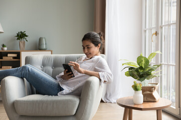 Cheerful relaxed young Indian woman using mobile phone, resting in cozy soft armchair in modern home Interior, shopping with online internet ecommerce app, touching screen, smiling