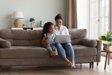 Happy pretty Indian mother showing learning app on laptop to little daughter. Mom and cute girl watching movie together, talking on video call, enjoying leisure in home living room cozy interior