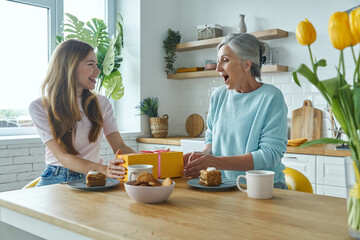Happy senior mother receiving a gift box from her adult daughter while both sitting at the kitchen