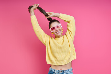 Happy young woman in funky glasses playing with her long hair against colored background