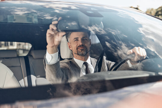 Confident man in formalwear adjusting rear view mirror while sitting on the front seat of a car - Powered by Adobe