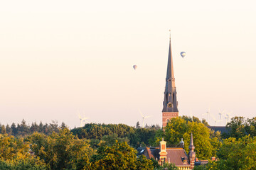 Fototapeta premium Cityscape small northern european city. European cathedral tower surrounded by green trees at sunset with hot air balloons. Breda, the netherlands