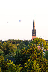 Fototapeta premium European cathedral tower surrounded by green trees at sunset with hot air balloons. Breda, the Netherlands