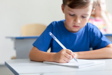 Naklejka na ściany i meble School child writing test on lesson. Pupil sitting at desk in classroom. Kid studying in elementary college. Primary student back to school. Education for children. Boy wearing in blue casual t-shirt