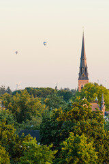 Fototapeta premium European cathedral tower surrounded by green trees at sunset with hot air balloons. Breda, the Netherlands