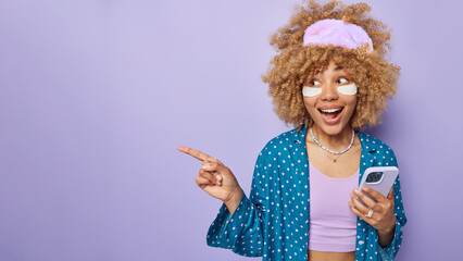 Cheerful surprised curly haired woman points away on blank space holds mobile phone sends text messages wears sleepmask and pajama shows empty place for your advertisement gives recommendations