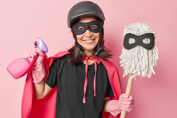Positive Asian woman dressed in superhero costume with cloak holds spray detergent and mop ready...