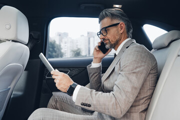 Confident mature businessman using digital tablet and talking on phone while sitting in the car