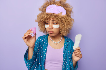 Unhappy frustated woman chooses between menstruation cup and sanitary napkin has critical days dressed in nightwear applies beauty patches under eyes to reduce puffiness isolated over purple wall