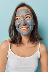 Vertical shot of dark haired woman smiles toothily applies beauty facial mask to remove pores dressed in casual t shirt isolated over blue background. Skin balancing and rejuvenation concept