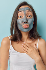 Photo of young Asian dark haired woman keeps lips folded applies clay mud mask keeps hand on chest dressed in casual t shirt isolated over blue background improves skin texture and clear acne