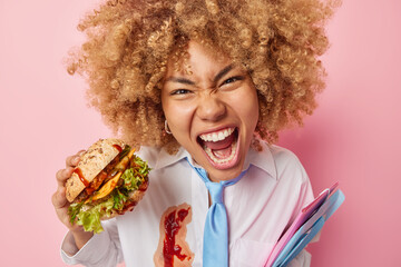 Outraged curly haired European woman feels angry and hungry eats appetizing burger holds folders...