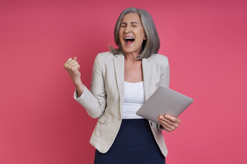 Happy senior woman holding digital tablet and gesturing while standing against pink background