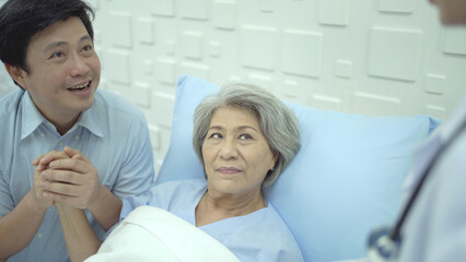 Senior Asian female patient resting on the medical bed in hospital and talking to her son. 