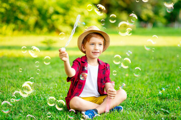 a little red-haired boy inflates soap bubbles on a green lawn in a hat and a red shirt in summer having fun playing