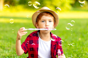 a little red-haired boy inflates soap bubbles on a green lawn in a hat and a red shirt in summer having fun playing