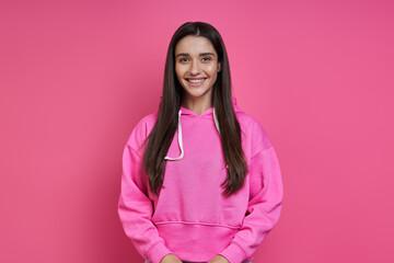 Beautiful young woman in hooded shirt looking at camera and smiling against pink background