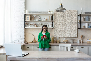 Beautiful woman in glasses and green shirt using red mobile phone at home in kitchen, smiling and happy