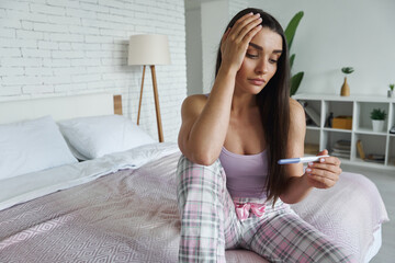 Frustrated young woman holding pregnancy test and touching head while sitting on bed at home