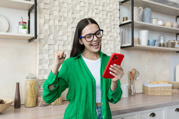 Beautiful young woman in glasses and green shirt read good news from red phone, brunette at home in...