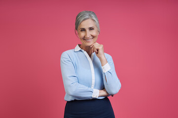 Confident senior woman holding hand on chin while standing against pink background