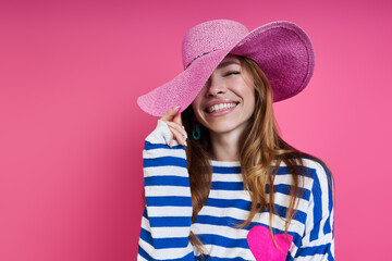Beautiful young woman adjusting her hat and smiling while standing against colored background