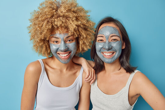 Photo of mixed race women apply beauty facial masks takes care of skin smile gladfully stand closely to each other dressed in t shirts isolated over blue background. Cosmetology and wellness - Powered by Adobe