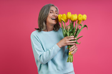 Happy senior woman smelling a bunch of tulips while standing against pink background