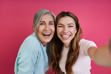 Happy senior mother and her adult daughter making selfie against pink background
