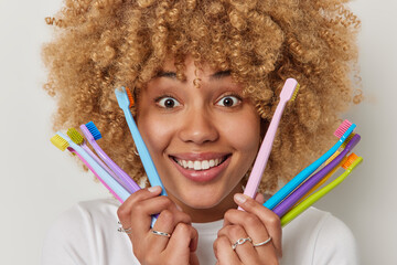 Headshot of positive glad woman with curly hair holds different variety of toothbrushes has well cared teeth takes care of oral hygiene isolated over white background. Morning routine concept