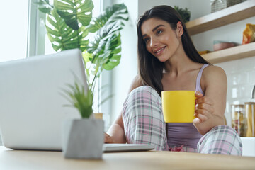 Beautiful young woman enjoying morning coffee and using laptop at the domestic kitchen