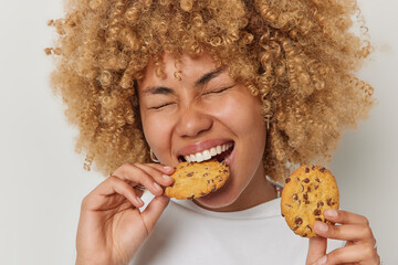 Close up shot of blonde curly haired woman eats cookies with chocolate bites deicious snack keeps eyes closed has sweet tooth dressed casually isolated over white background. Unhealthy eating - 520365849