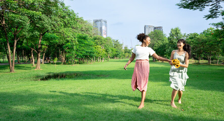 Fototapeta premium Friend girl concept.Two smiling girls run at green grass at background of blue sky with clouds.Young adult African Asian American woman walk on summer field. Empty space for inscription or objects.