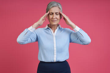 Senior woman suffering from headache while standing against pink background